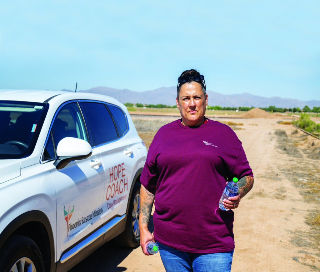 Manette holds a water bottle and looks at the camera standing next to a Phoenix Rescue Mission Hope Coach in the desert. 