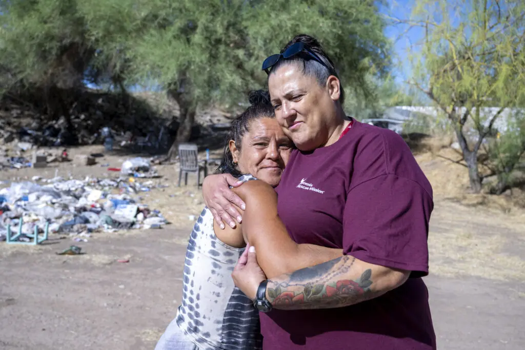 Manette hugs a homeless woman in front of an encampment in the desert sun, reaching out on behalf of PRM.