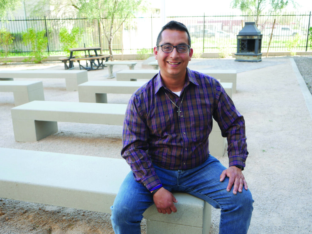 Robert Leon sits on a park bench in the shade, smiling at the camera. 