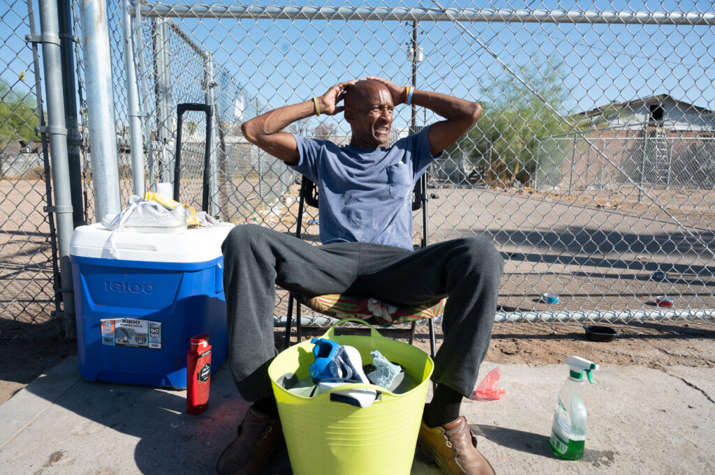 A man on the streets grimaces in the heat, seated in direct sunlight.